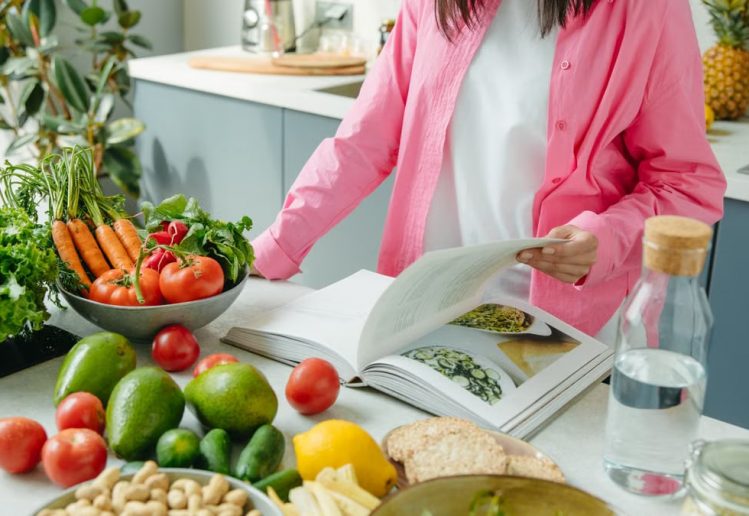 A person browsing a cookbook surrounded by fresh vegetables in a bright kitchen setting.