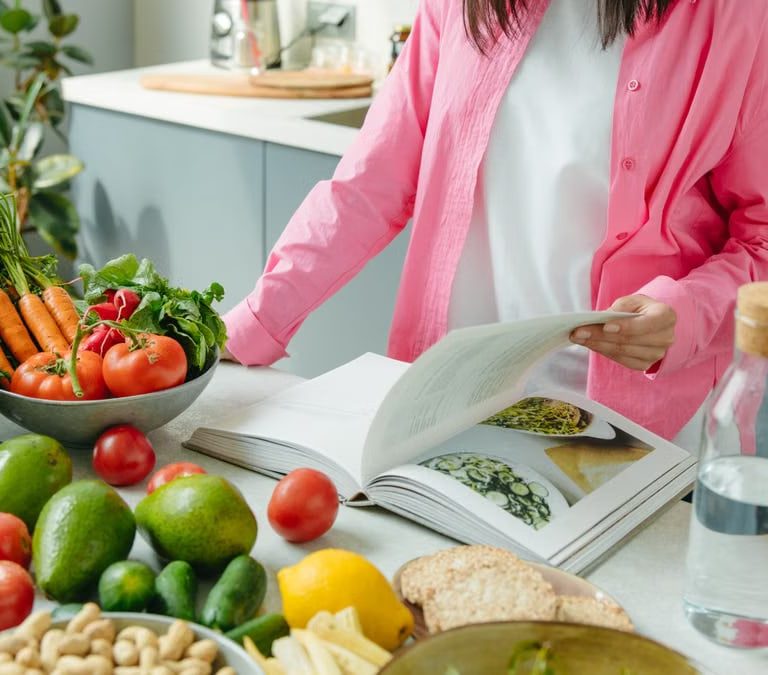 A person browsing a cookbook surrounded by fresh vegetables in a bright kitchen setting.