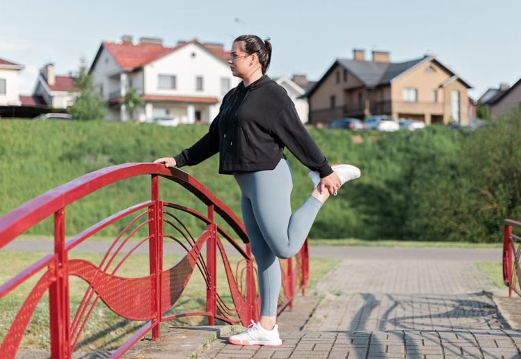 A woman in active wear performs a stretching exercise on a red bridge in a suburban park setting.